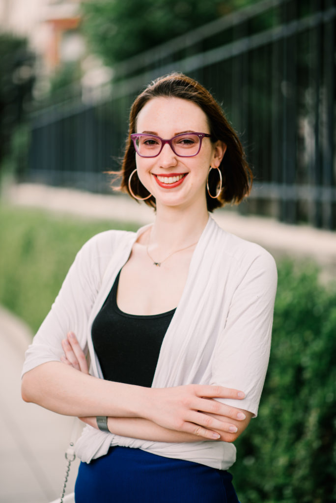 A young woman wearing glasses and professional clothes poses for the camera with a smile, arms crossed.