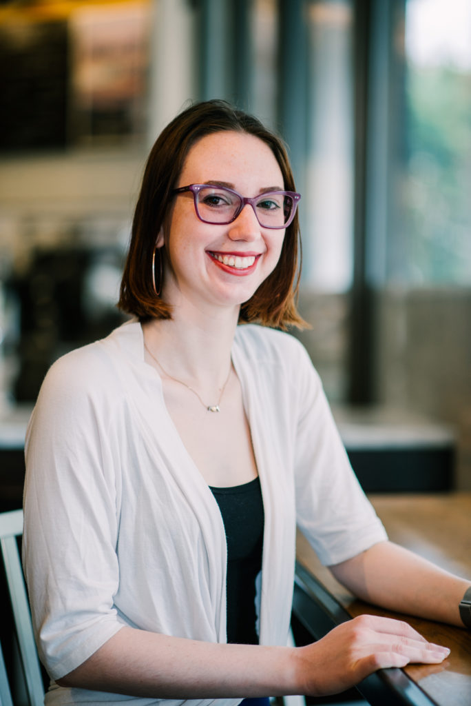 A young woman wearing glasses and professional clothes in a cafe smiles at the camera.